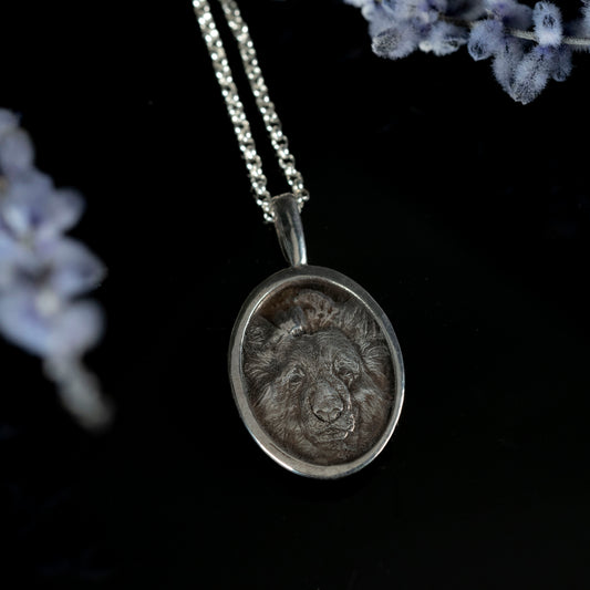 Silver pendant with dimensional engraving of dog design on a chain against a dark background with flowers.