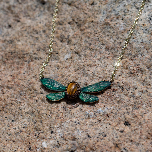 A gold chain necklace with an insect pendant featuring green wings and a Tiger Eye center, displayed on a speckled stone surface.