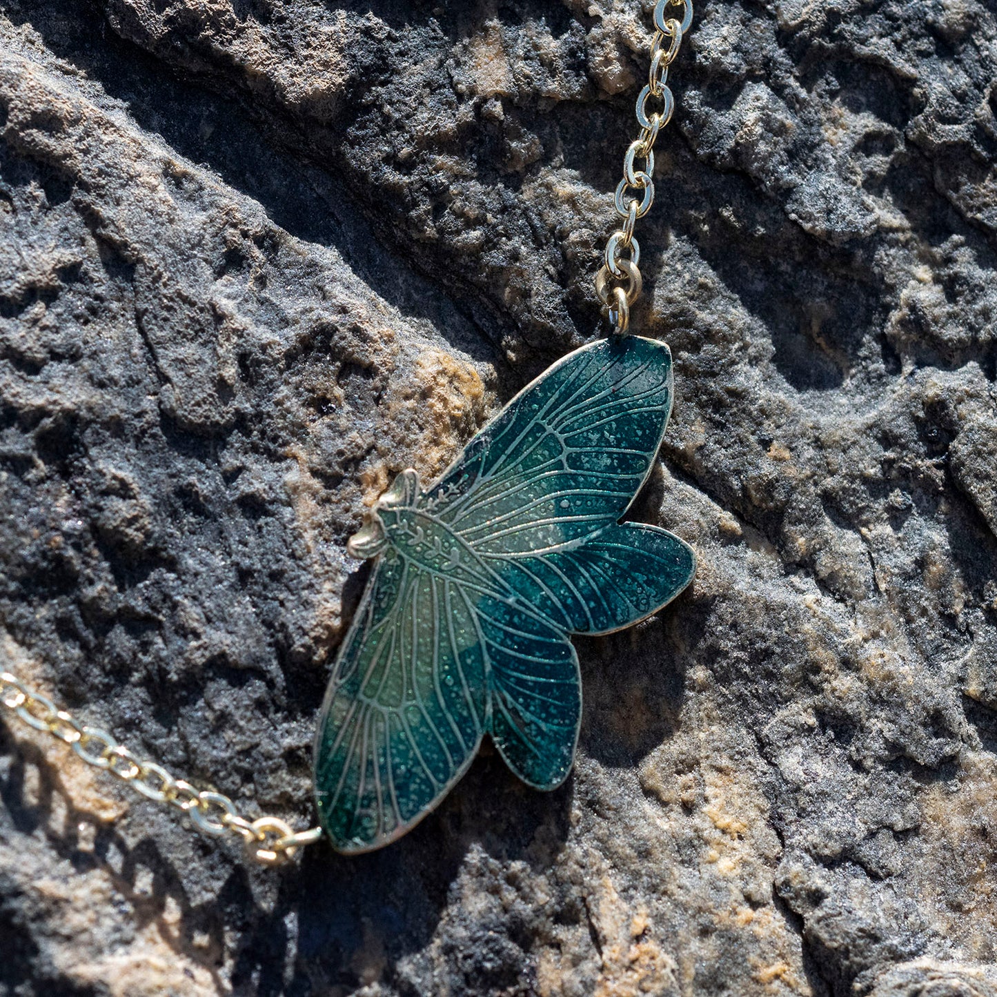A gold-toned moth pendant necklace with a dark teal enamel fill, resting on a textured dark gray rock surface.