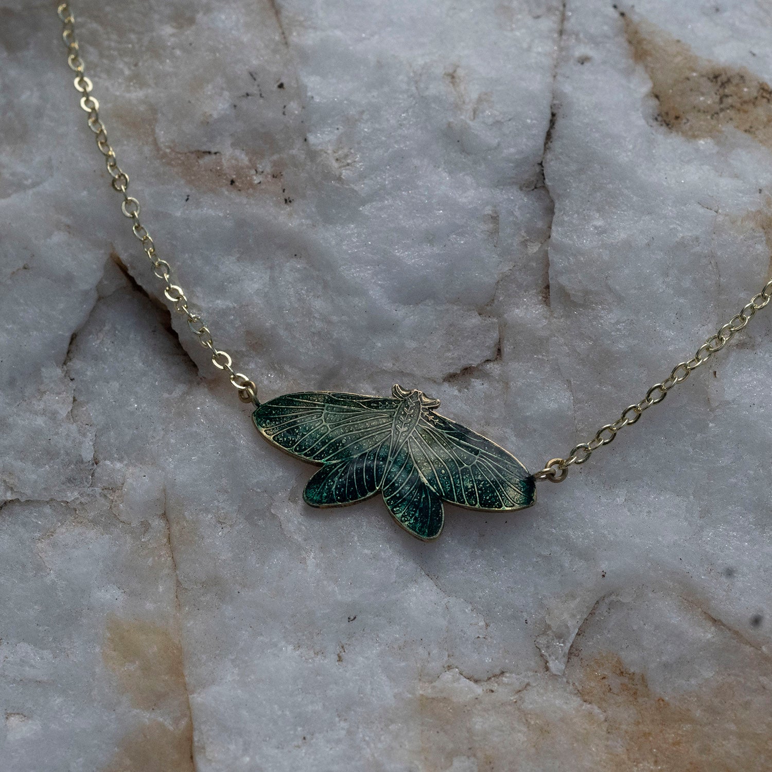 A delicate gold chain necklace featuring an intricately detailed moth pendant with dark green and gold enamel wings, resting on a textured white and gray stone surface.