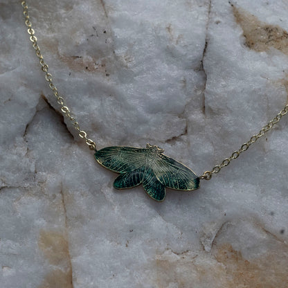 A delicate gold chain necklace featuring an intricately detailed moth pendant with dark green and gold enamel wings, resting on a textured white and gray stone surface.