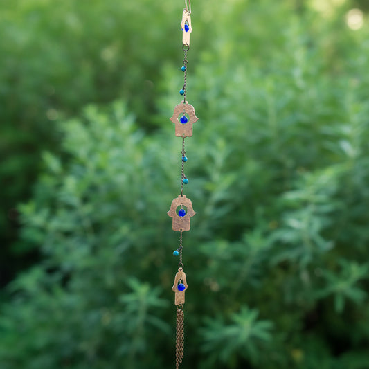 A hanging Hamsa wall hanging ornament with four copper hamsa charms, each featuring a Lapis blue evil eye gemstone, suspended from a chain with Turquoise beads, set against a blurred wooden fence and green plants.