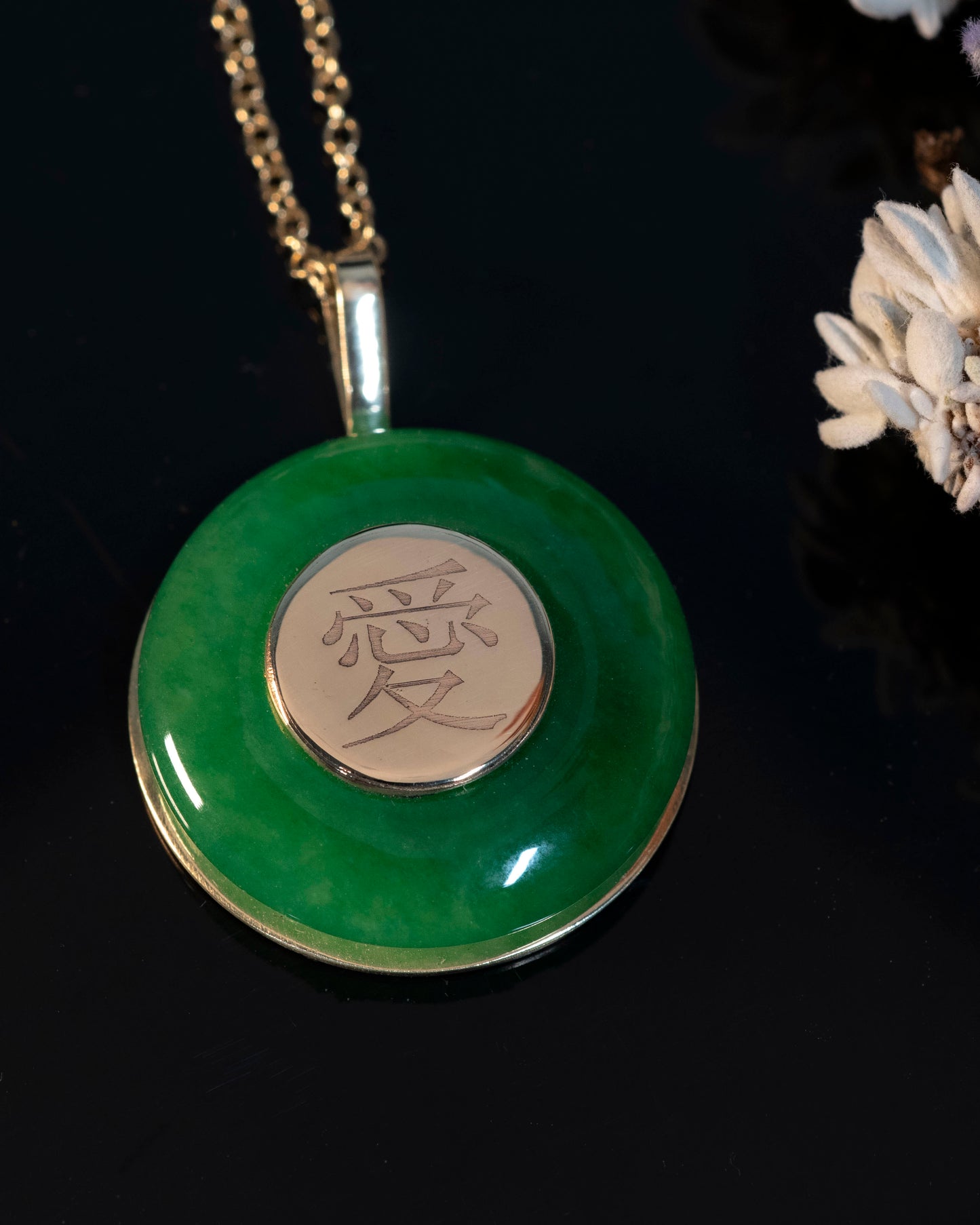 A round green jade pendant with a traditional Chinese symbol for love engraved on it, suspended from a gold chain.