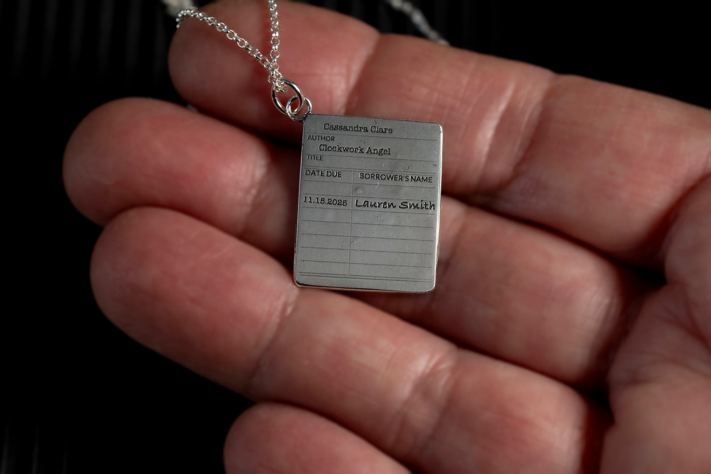 Silver library card pendant with engraved text held in a hand against a dark background