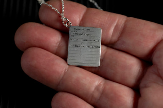 Silver library card pendant with engraved text held in a hand against a dark background
