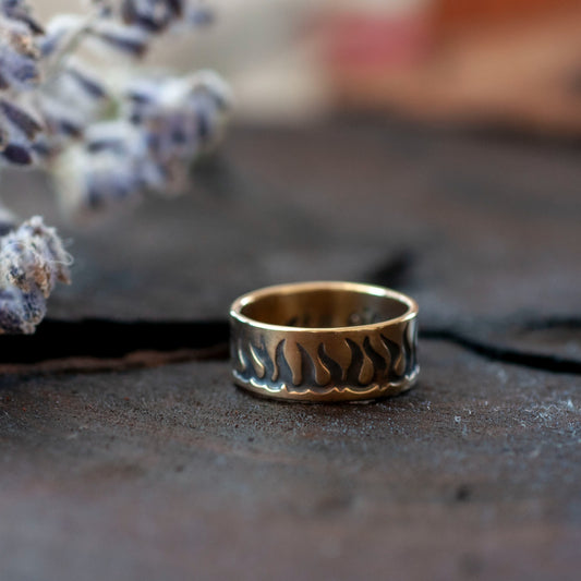 A gold wedding band with an fire engraved pattern, displayed on a textured surface with dried flowers in the background.