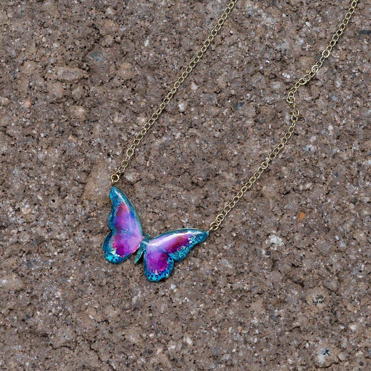 A gold-tone chain necklace with a butterfly pendant featuring vibrant purple and pink wings with teal-blue edges, displayed on a textured brown stone surface.
