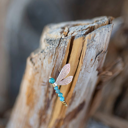 A dragonfly-shaped earring with a textured rose gold body and translucent wings, accented with blue beads and gold spacer beads, resting on a piece of weathered driftwood.