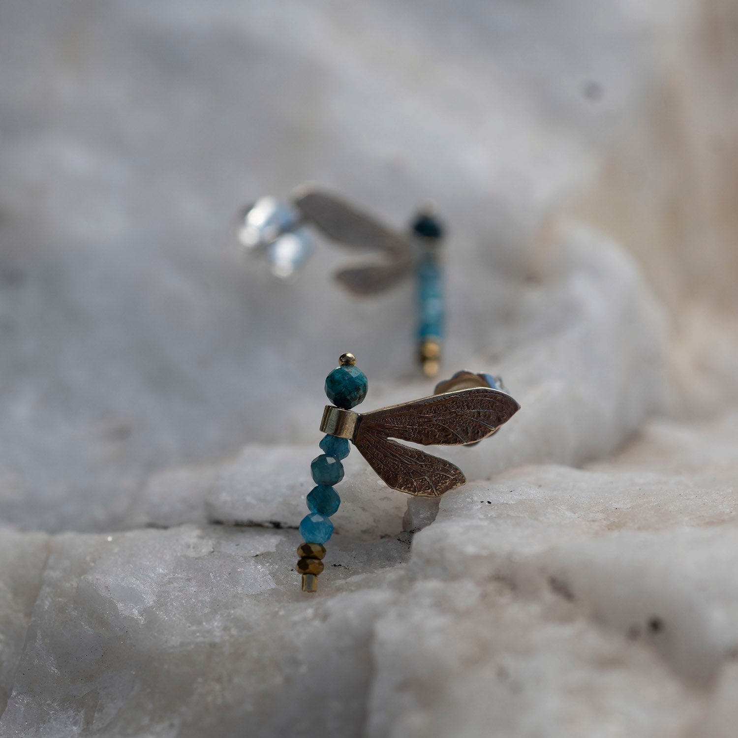 A pair of dragonfly earrings resting on a textured white stone surface. The front earring is in sharp focus, featuring a brown wing, a gold post, and a vertical bead strand with turquoise blue beads and a gold spacer.